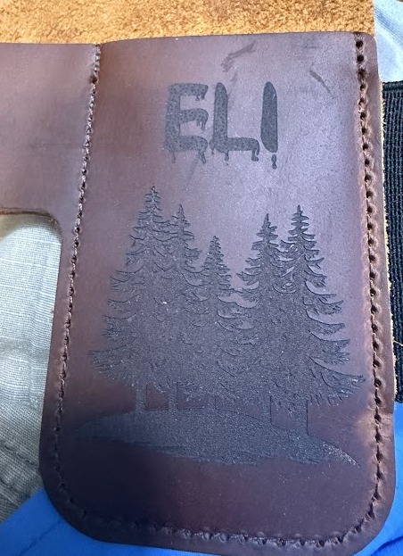 Brown leather knife case engraved with the name ELI and a pine forest scene