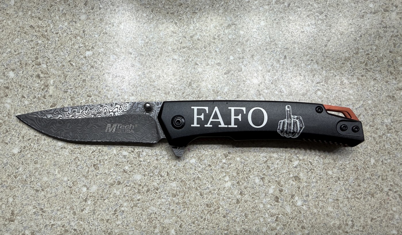 Black folding knife with damascus blade, handle engraved FAFO with a hand gesture graphic, shown open